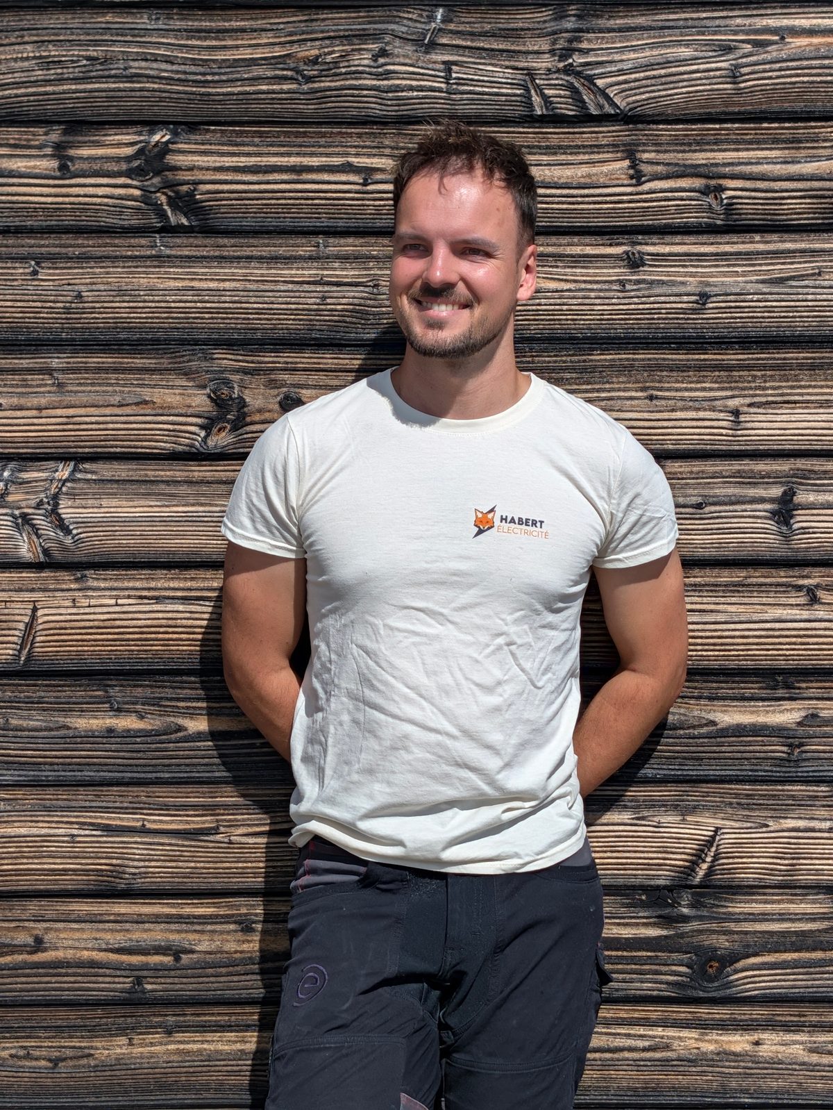 Théo Habert devant un mur en bois, portant un t-shirt Habert Électricité